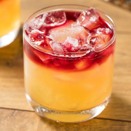 A cocktail with a yellow base, topped with red liquid and ice cubes in a short glass sitting on a wooden surface.