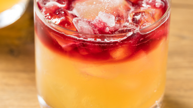 A cocktail with a yellow base, topped with red liquid and ice cubes in a short glass sitting on a wooden surface.