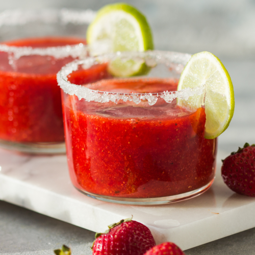Two strawberry margaritas with lime slices and salt rims are on a marble tray, surrounded by fresh strawberries and limes.