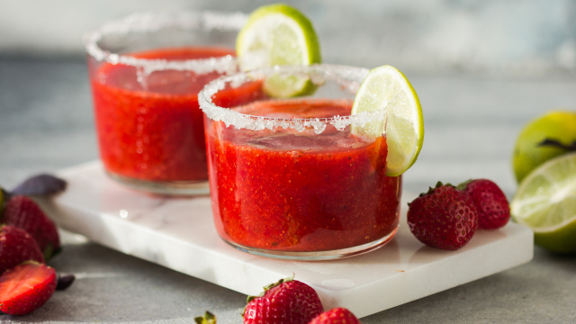 Two strawberry margaritas with lime slices and salt rims are on a marble tray, surrounded by fresh strawberries and limes.