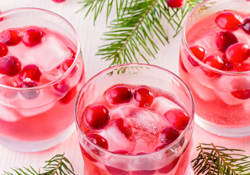 Three glasses of a pink drink with ice and cranberries, surrounded by pine branches on a white surface.