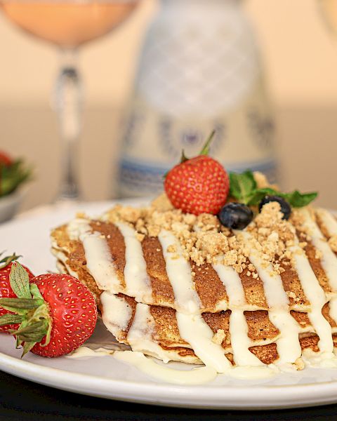 A frosted cake with white drizzle sits on a plate, garnished with strawberries and a berry on top; a bowl of strawberries and drinks in the background.