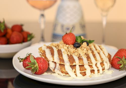 A frosted cake with white drizzle sits on a plate, garnished with strawberries and a berry on top; a bowl of strawberries and drinks in the background.