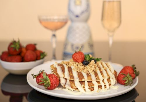 A plate of pancakes topped with strawberries, cream, and nuts, a bowl of strawberries, and two glasses, one with a beverage, on a reflective table.