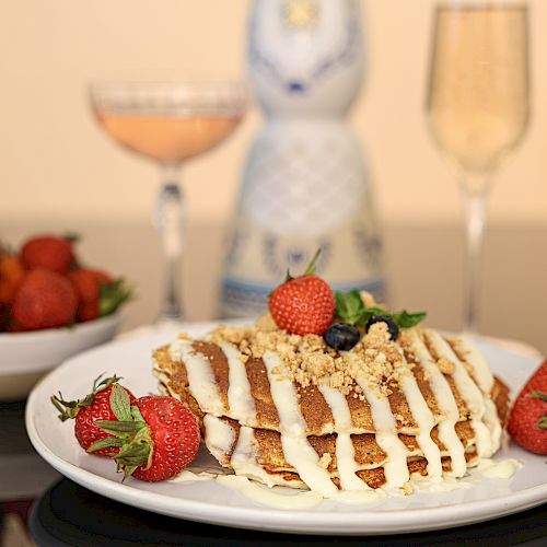 A plate of pancakes topped with strawberries, cream, and nuts, a bowl of strawberries, and two glasses, one with a beverage, on a reflective table.