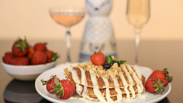 A plate of pancakes topped with strawberries, cream, and nuts, a bowl of strawberries, and two glasses, one with a beverage, on a reflective table.