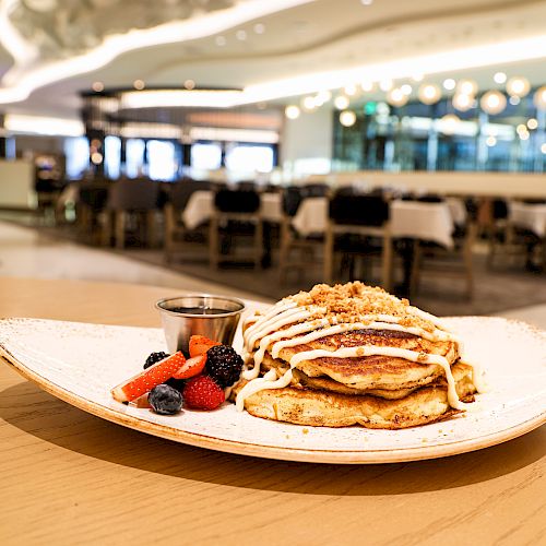 A plate of pancakes topped with cream, accompanied by berries and syrup, is in focus, with a restaurant setting in the background.