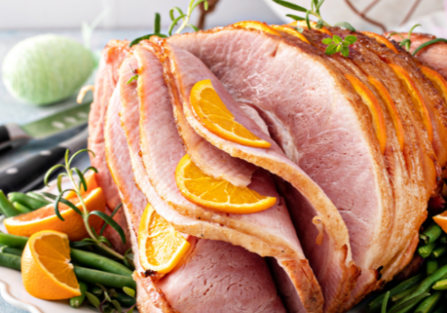 A sliced ham garnished with orange slices and herbs is presented on a platter with green beans, creating a festive meal setting.