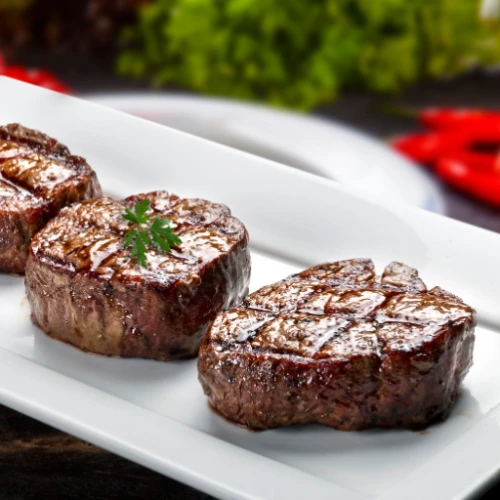 Three grilled steaks on a white rectangular plate, garnished with a small sprig of parsley, with a blurred background.