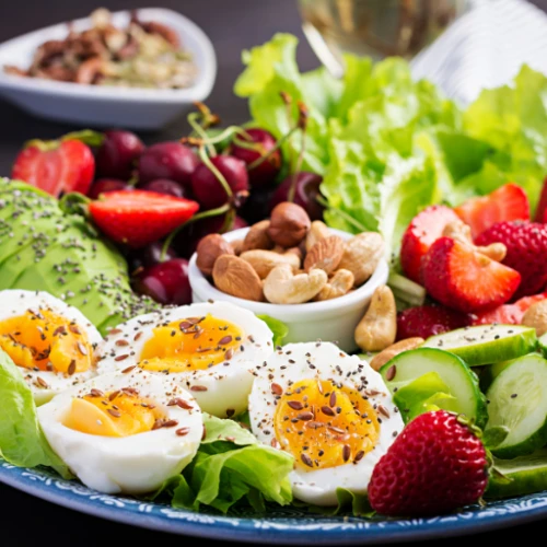 A vibrant salad with soft-boiled eggs, nuts, strawberries, cucumbers, avocado slices, cherries, and leafy greens, garnished with seeds.