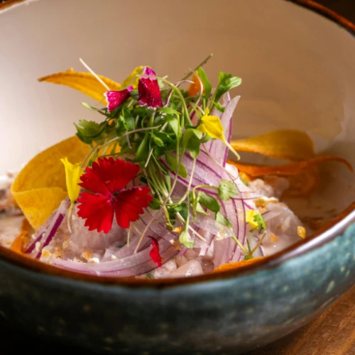 A bowl of ceviche topped with sliced red onions, edible flowers, microgreens, and plantain chips.