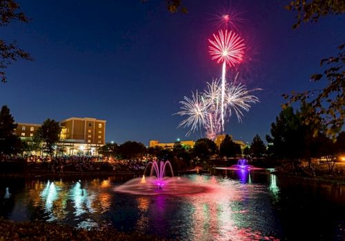 A vibrant fireworks display lights up the night sky over a pond with fountains, surrounded by buildings and trees.