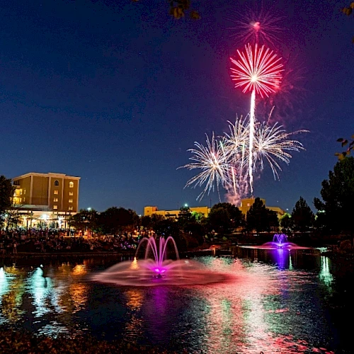 A vibrant fireworks display lights up the night sky over a pond with fountains, surrounded by buildings and trees.