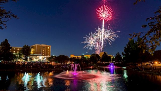 A vibrant fireworks display lights up the night sky over a pond with fountains, surrounded by buildings and trees.