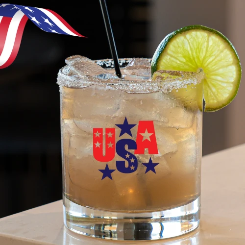 A cocktail glass with a lime slice and straw, featuring a "USA" design with stars. There's also a wavy American flag ribbon nearby.