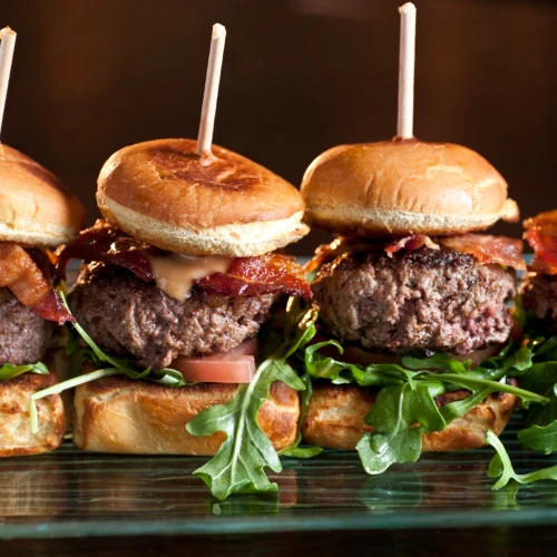 Four sliders with beef patties, bacon, and arugula on buns, held together with skewers, on a glass surface.