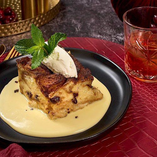 A dessert on a black plate with custard, topped with mint and cream, sits by a glass of amber liquid on a red textured mat.