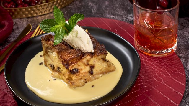 A dessert on a black plate with custard, topped with mint and cream, sits by a glass of amber liquid on a red textured mat.
