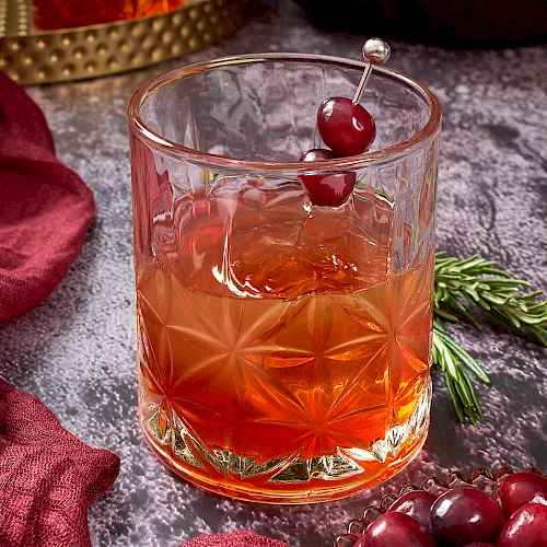A glass with an amber cocktail garnished with cranberries on a skewer, surrounded by cranberries, rosemary, and red fabric.