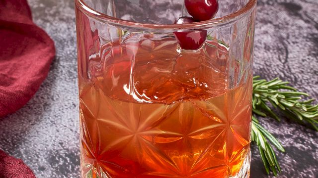 A glass with an amber cocktail garnished with cranberries on a skewer, surrounded by cranberries, rosemary, and red fabric.