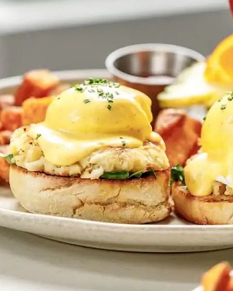 Two yolky Eggs Benedict-style mini sandwiches with hollandaise, plus home fries on a plate and a citrus garnish, all brunchy delicious.