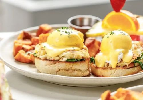 Two yolky Eggs Benedict-style mini sandwiches with hollandaise, plus home fries on a plate and a citrus garnish, all brunchy delicious.