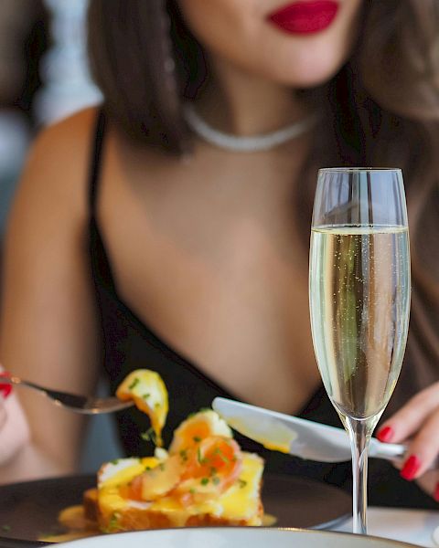 A woman in a black dress with red lipstick enjoys a fancy meal, with a champagne flute and a plated dessert on the table.