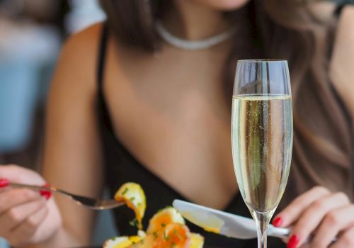 A woman in a black dress with red lipstick enjoys a fancy meal, with a champagne flute and a plated dessert on the table.