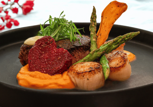 A plated beef tenderloin with orange puree, seared scallops, asparagus, a heart-shaped beet slice, and crispy root vegetables on a dark plate.