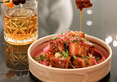 A glass of whiskey on the rocks beside a bowl of glazed meat bites on a wooden plate, being skewered, tempting and glossy.