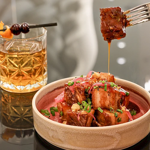 A glass of whiskey on the rocks beside a bowl of glazed meat bites on a wooden plate, being skewered, tempting and glossy.