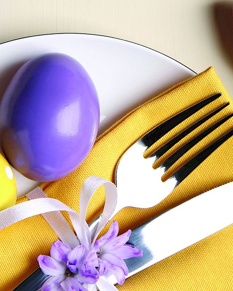 Colorful Easter table setting with a yellow napkin, white plate, purple and yellow eggs, a white-handled fork and knife, ribbons, and tulips.