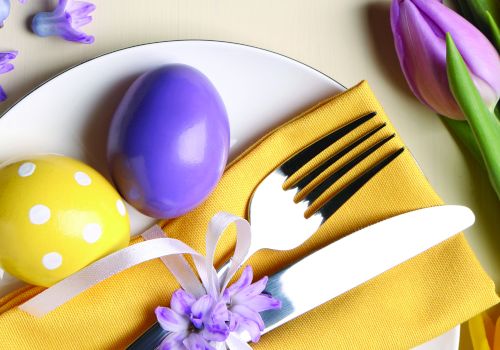 Colorful Easter table setting with a yellow napkin, white plate, purple and yellow eggs, a white-handled fork and knife, ribbons, and tulips.