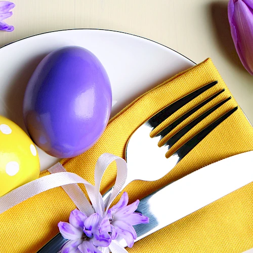 Colorful Easter table setting with a yellow napkin, white plate, purple and yellow eggs, a white-handled fork and knife, ribbons, and tulips.