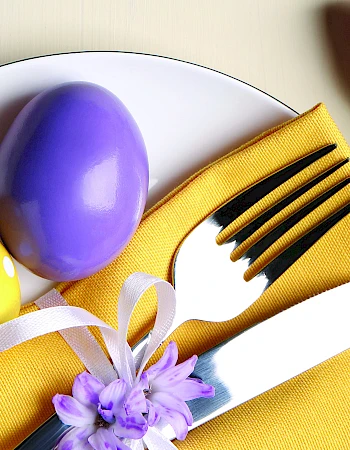 Colorful Easter table setting: purple egg, yellow polka-dotted egg, white plate with yellow napkin, silverware, purple flowers, and tulips.