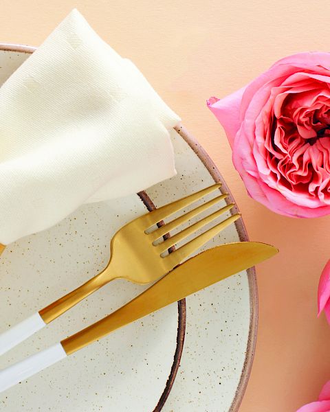 A elegant place setting with a white napkin folded over a plate, gold cutlery, surrounded by pink roses on a peach background.