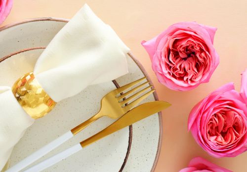 A elegant place setting with a white napkin folded over a plate, gold cutlery, surrounded by pink roses on a peach background.