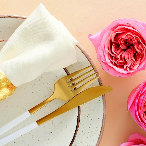 A elegant place setting with a white napkin folded over a plate, gold cutlery, surrounded by pink roses on a peach background.