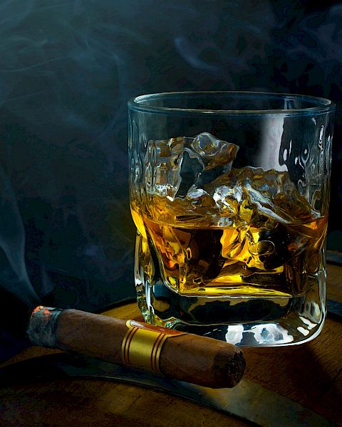 A glass of whisky with ice on a wooden surface, a lit cigar resting nearby, and wisps of smoke curling in a dark, moody setting.