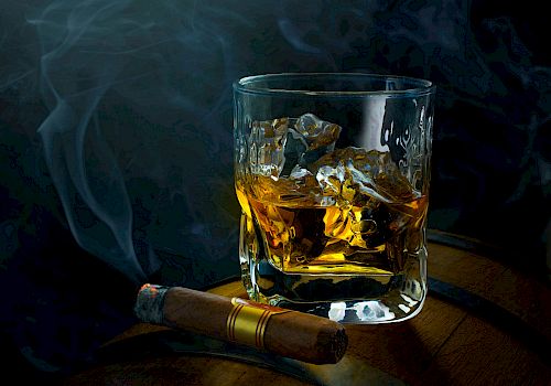 A glass of whisky with ice on a wooden surface, a lit cigar resting nearby, and wisps of smoke curling in a dark, moody setting.