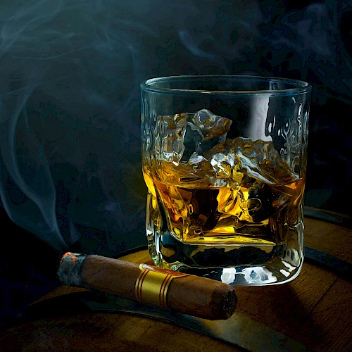A glass of whisky with ice on a wooden surface, a lit cigar resting nearby, and wisps of smoke curling in a dark, moody setting.