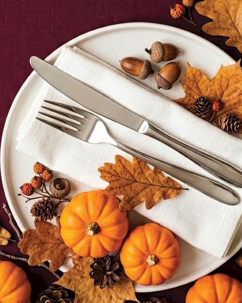 A white plate with fork and knife, surrounded by autumn leaves, acorns, pinecones, and orange pumpkins on a maroon tablecloth.