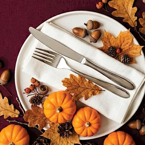 A white plate with fork and knife, surrounded by autumn leaves, acorns, pinecones, and orange pumpkins on a maroon tablecloth.